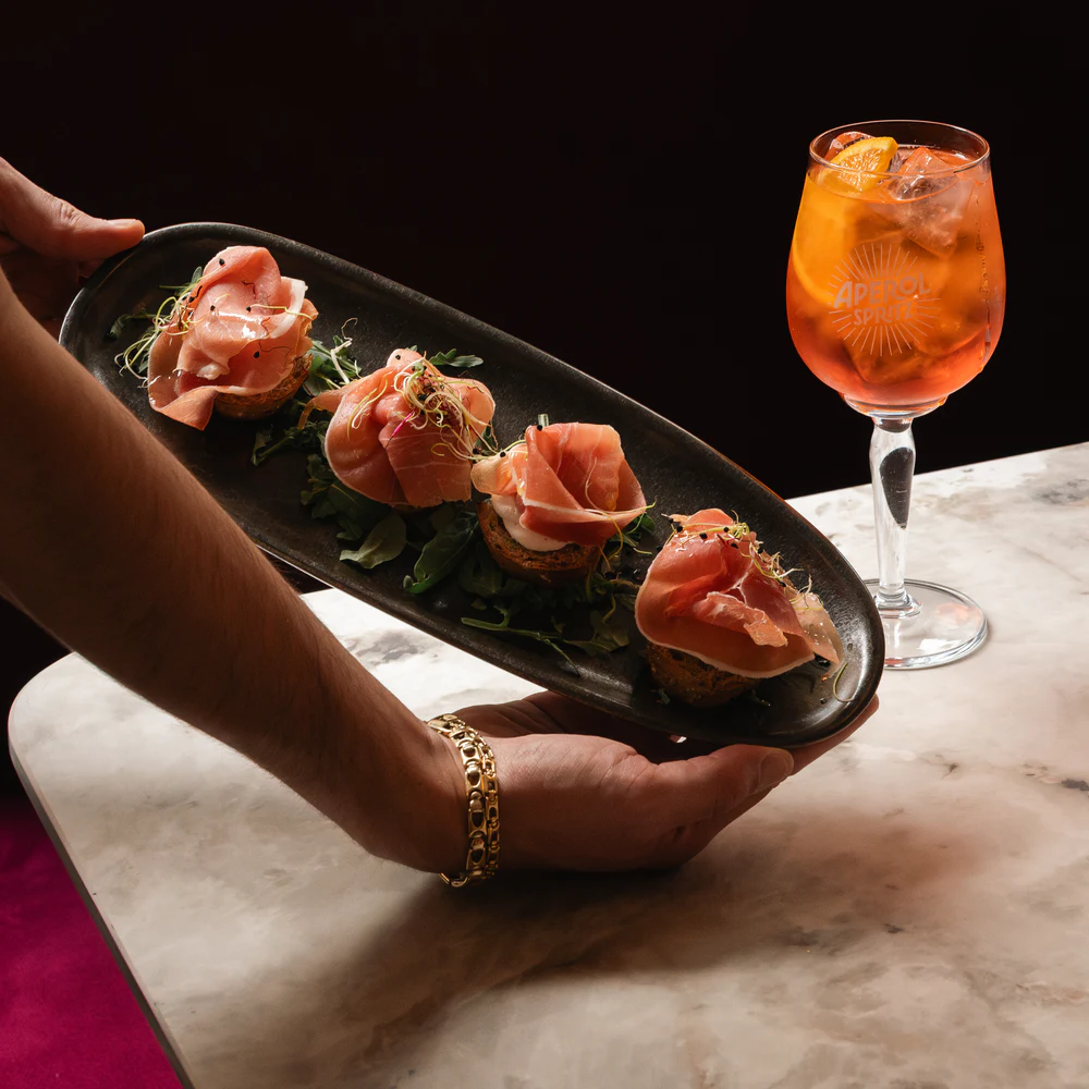 A board of crostini carried past an Aperol spritz on a marble bar.