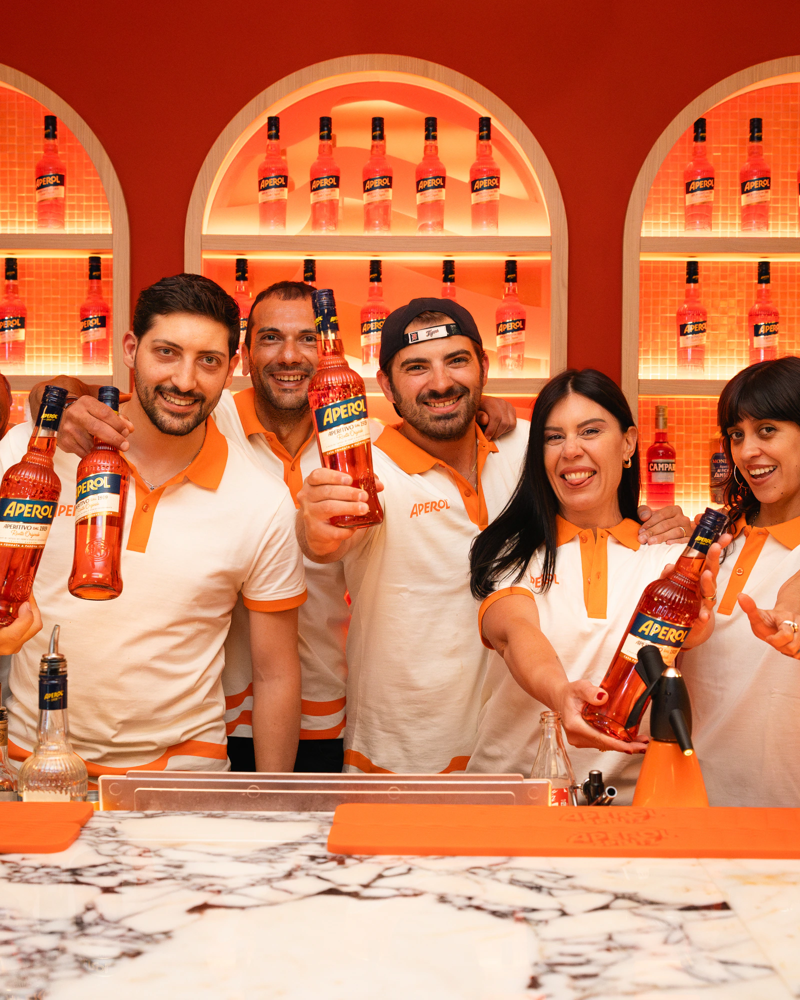 The Aperol team behind the bar, bottles raised, under the lit bottle wall